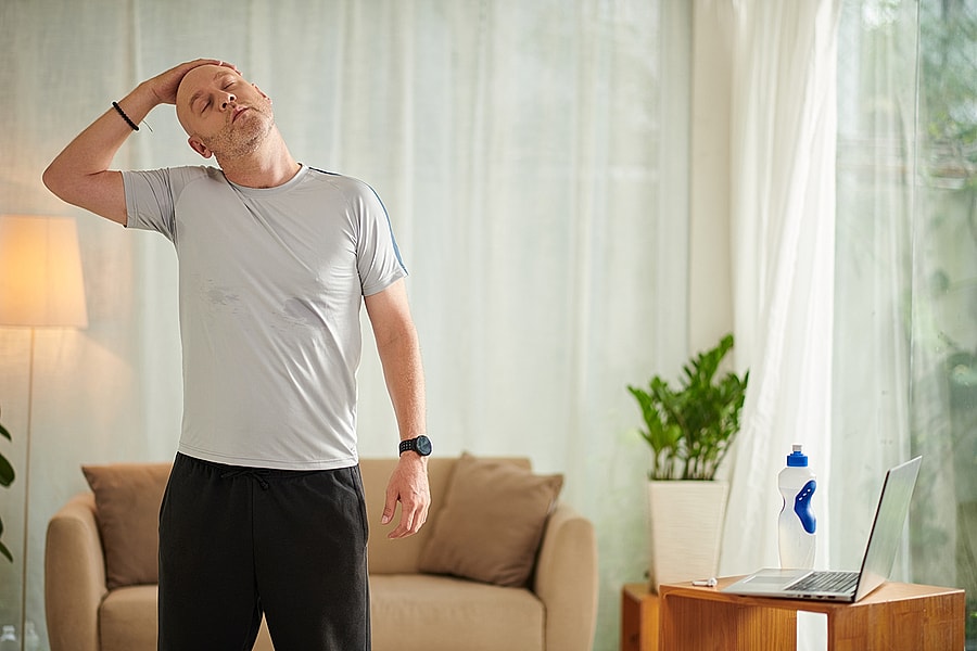 Man stretching his neck at home near laptop and water bottle.