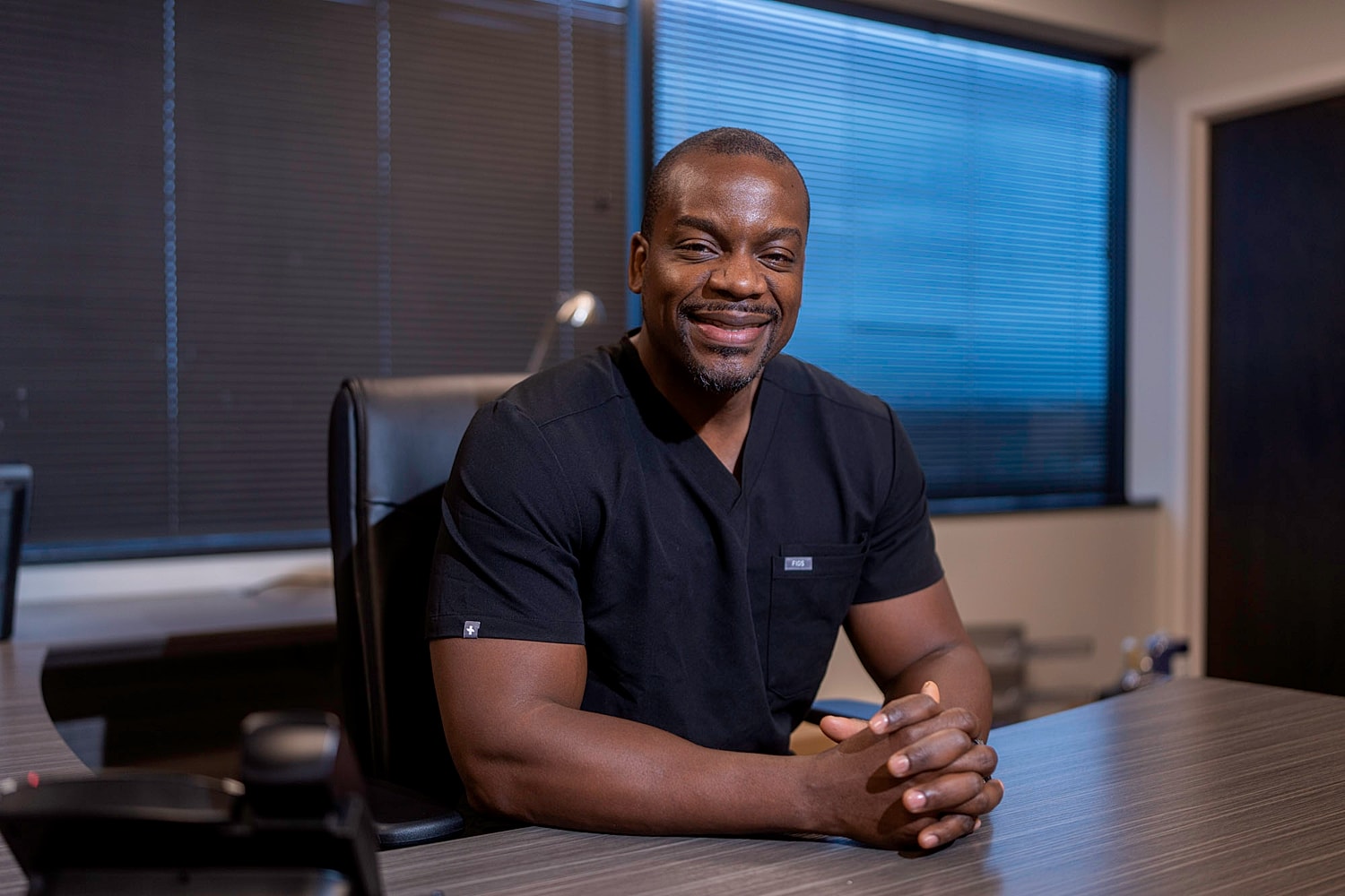 Smiling spine surgeon in black scrubs at desk.